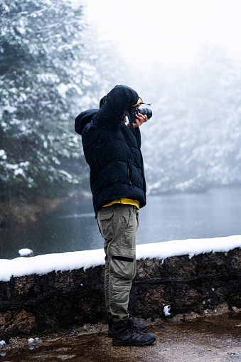 Hispanic explorer in snowy forest captures white pine trees surrounding frozen lake.