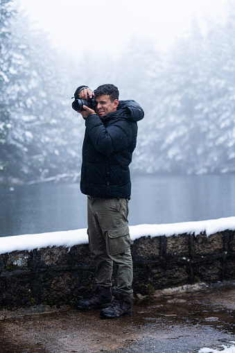 Latinx young man in black coat captures snowy forest lake with professional camera.