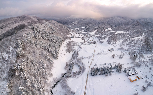 Drone view above a picturesque small village located in a valley at the feet of a wooded hill, along a stream. Winter season, every house and field and tree is covered in snow. Carpathia, Romania.