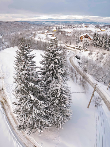 Drone view above a picturesque small village located in a valley. A road winds along coniferous trees. Winter season, every house and field and tree is covered in snow. Carpathia, Romania.