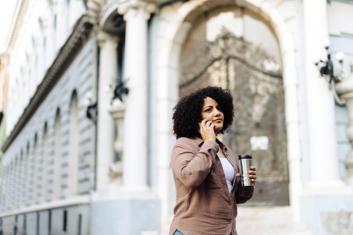 Young afro woman drinking coffee and using her smart phone on the street