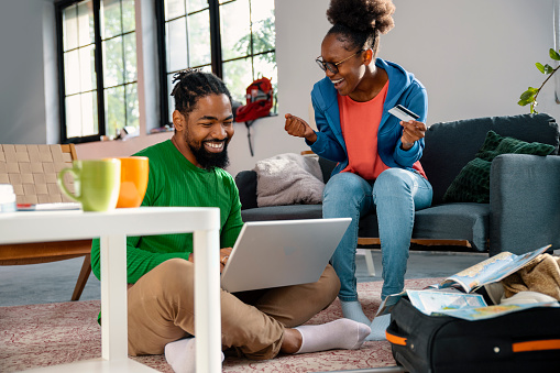 Happy black couple of travelers make a trip reservation buying online with credit card. Couple using laptop in their living room to make travel reservation