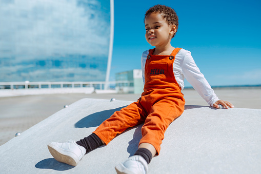 Cute african baby toddler in a orange overalls sits on a stone bench by the sea at city. Happy girl playing in summer alone.