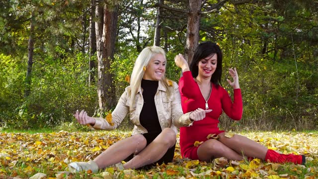 Two Female Friends Throwing Autumn Leaves On The Grass In A Park
