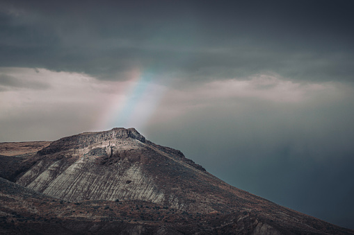 A rainbow cuts through thick dark clouds to illuminate the top of a solitary mountain.