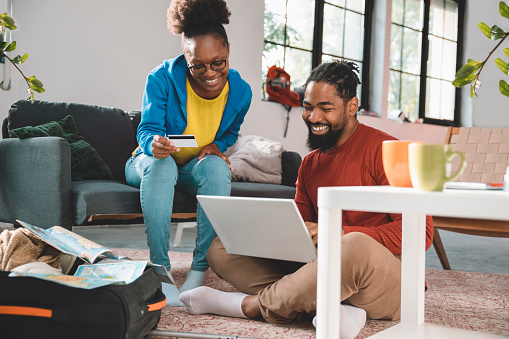 Happy black couple of travellers make a trip reservation buying online with credit card. Couple using laptop in their living room to make travel reservation