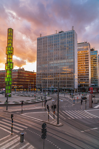 Stockholm, Sweden - April 10th 2024: Square of Sergels torg and the Hötorg skyscrapers of Stockholm at sunset, orange and yellow sky, clouds. The main monument lit up in green.