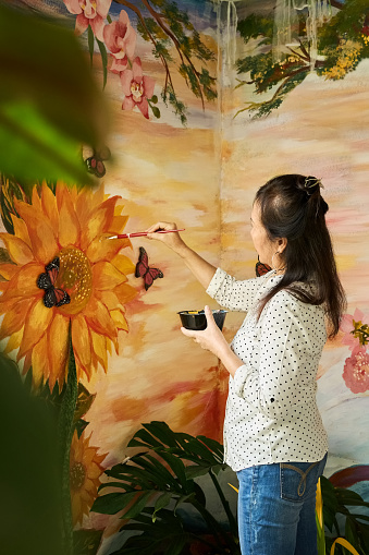 Senior asian woman painting floral wall mural on backyard wall of her house