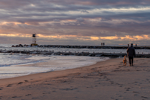 Avon By the Sea, New Jersey, USA - January 21, 2023 - Person  and their  dog walking along the beach at Avon by the Sea by the Shark River inlet at sunrise.