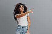 Shocked amazed afro-haired Latin woman wearing casual clothes standing pointing index finger aside on mock up copy space put hand on leg isolated on gray blue colour background, studio portrait