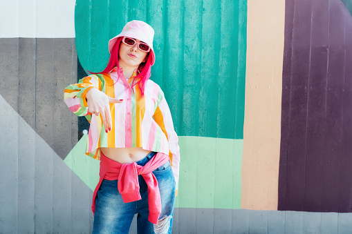 Young woman with pink hair and sunglasses in Bucket hat and multicolor strippled shirt posing on graffiti wall background. Street artist with mural, hipster, blogger. Urban street fashion.