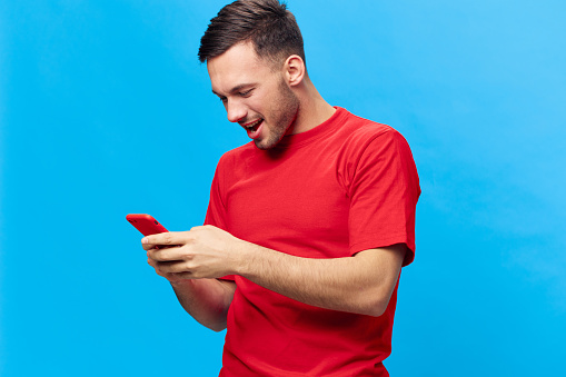 Concentrated happy smiling tanned handsome man in red t-shirt chatting with friends using phone posing isolated on blue studio background. Copy space Banner Mockup. Online People lifestyle concept