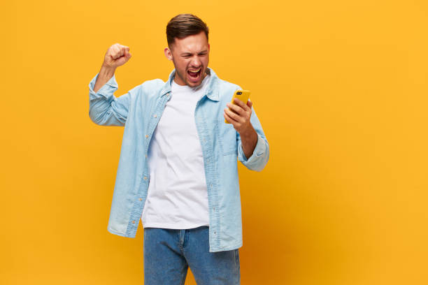 istock Angry irritated tanned handsome man in blue basic t-shirt scream at smartphone posing isolated on orange yellow studio background. Copy space Banner Mockup. Online Social media concept. Bad news 2148747872