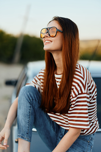 A fashionable woman smiles sweetly in stylish sunglasses, a striped t-shirt and jeans, sits on the trunk of a car and looks at the beautiful nature of autumn. Travel lifestyle. High quality photo