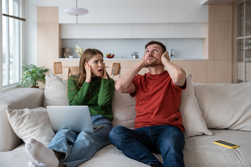 Exhausted irritated couple sitting on sofa with ears closed because of unpleasant nervous sound, knock from neighboring flat. Disadvantages of neighborhood, living in residential apartment buildings