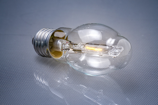 incandescent bulb on a reflective surface. The bulb is turned on and the filament inside is glowing. It has a clear glass envelope allowing the internal parts to be visible