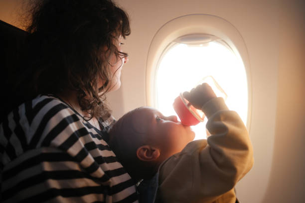 A little boy is drinking water while he's sitting on a plane with his mother. The mother is looking outside the window.