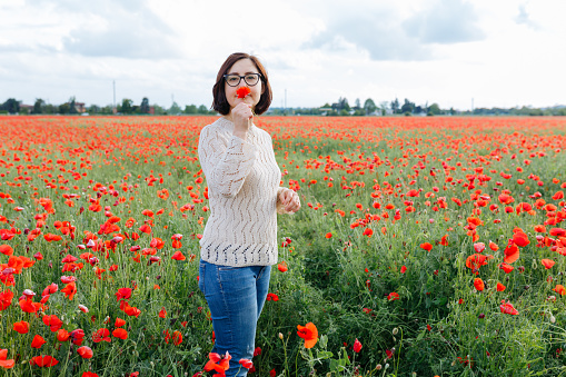 dark haired woman standing in the poppy field holding flower in her hand looking at camera