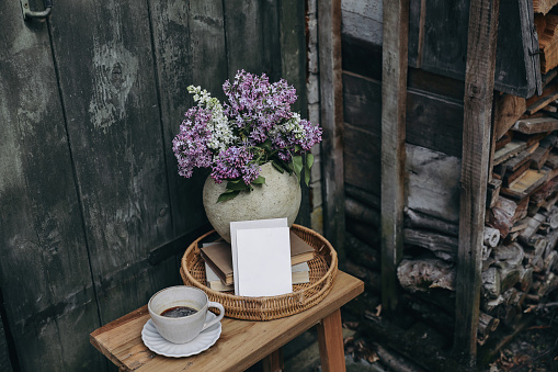 Moody rustic spring outdoor still life. Purple, white lilac flowers bouquet in textured vase. Cup of coffee, books. Greeting card, invitation mockup, blurred old wooden door background. Pile of firewood.