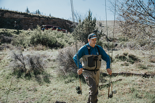 A man carries multiple fishing rods to the river while on a fly fishing trip with friends in Central Oregon on a sunny Spring day.