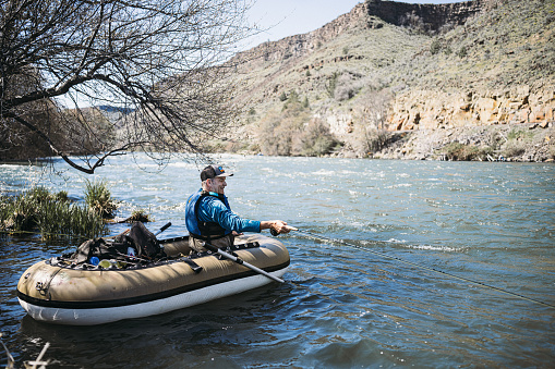 A healthy and active Caucasian man sits in an inflatable boat while fly fishing in the Deschutes River during a weekend trip with his friends.
