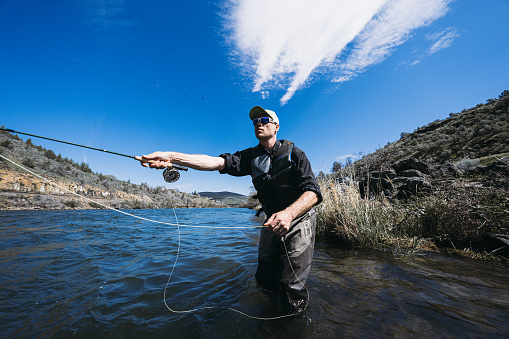 An active middle aged Caucasian man uses a rod to catch fish while standing knee deep in the Deschutes River on a Spring day.