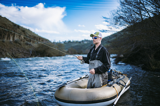 A Caucasian man fly fishes from a single person inflatable boat in the Deschutes River while on a fishing trip with friends in Central Oregon.