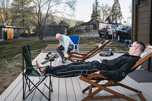A retired senior Caucasian man sits on a chair on the deck of his vacation home in Central Oregon and takes off his waders, after spending the day fly fishing with his adult son and friends along the Deschutes River.