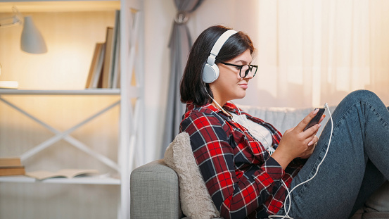 Mobile music. Song listening. Gadget leisure. Satisfied woman in headphones enjoying phone playlist on cozy couch at home interior.