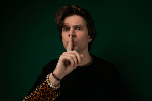 A man with a serious expression holding his index finger to his lips, signaling for silence. He is standing in a room with a neutral background.