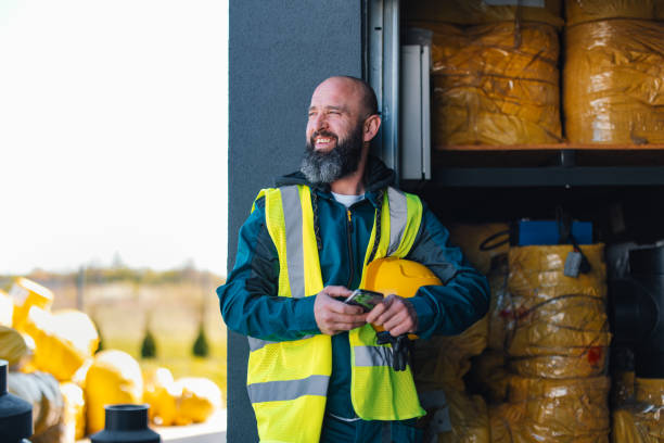 A cheerful bearded warehouse worker in a reflective safety vest stands by industrial storage, holding a tablet and hard hat. Captures the essence of safety and efficiency in industry settings.
