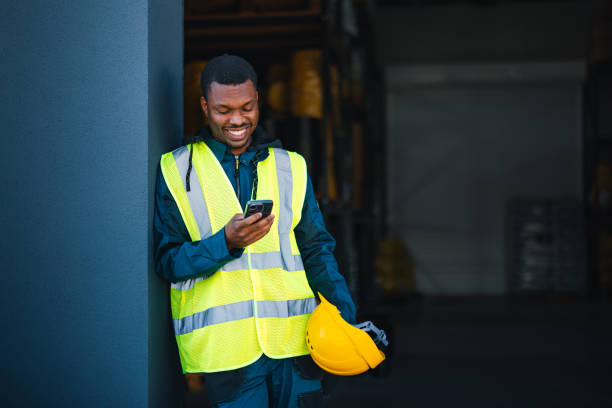 istock A male worker in a safety vest smiles while using a smartphone, standing in an industrial warehouse setting. He holds a yellow safety helmet, signaling a focus on work safety and communication. 2148771450