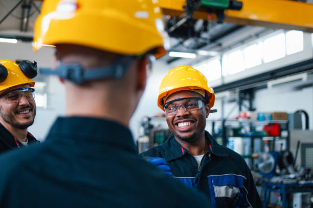 Three diverse workers wearing safety helmets smile and discuss in a busy factory setting, showcasing teamwork and industry safety. Smiling Workers Discussing at a Busy Factory Warehouse manufacturing occupation stock pictures, royalty-free photos & images