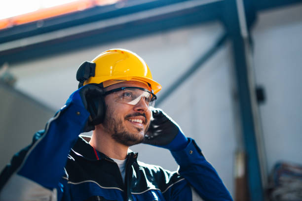 A cheerful male worker wearing a safety helmet and protective gear speaks on a headset in a bustling factory environment, emphasizing industry safety and productivity. Smiling Worker in Safety Helmet Communicating in Factory working class stock pictures, royalty-free photos & images