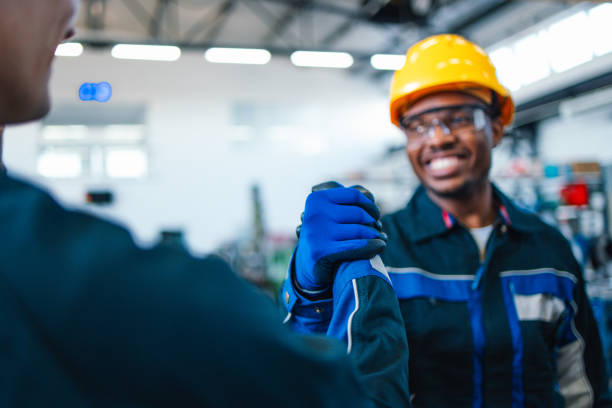 istock Two factory workers in a warehouse, one wearing a yellow safety helmet, share a friendly greeting with smiles. Focus on teamwork and safety in an industrial setting. 2148771936