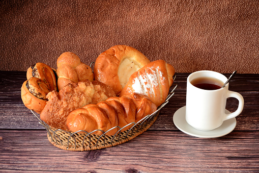 A cup of hot tea on a saucer and a wicker basket with an assortment of homemade fresh pastries on a wooden table. Close-up.
