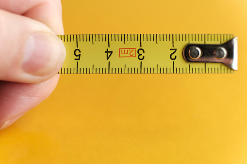 Metallic tape measure held by finger with orange background. It is a macro image and we can see few centimeters of the tape. Construction and work tool concept.