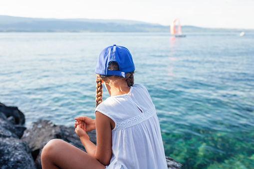 A young girl in a blue baseball cap and white dress enjoys a serene moment by the lake, watching sailboats glide across the water.