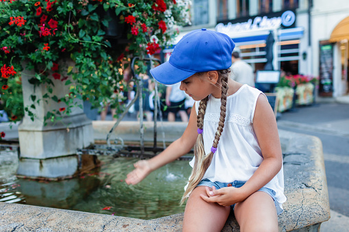A young girl in a blue cap sits by a stone fountain, gently touching the water. Vibrant flowers cascade from the fountain, adding color to an urban setting