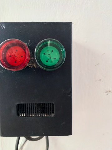 An intercom system affixed to a white wall. The intercom, encased in black, features a red button that shows signs of wear and dust, suggesting frequent use over the years. A second button is partially obscured by a green square, leaving its purpose to the imagination.
