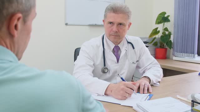 a doctor listening to an elderly patient and writing down the symptoms. An elderly man has an appointment with a specialist doctor at the clinic