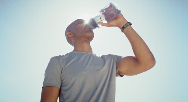 Black man, drinking water or break in fitness, training or workout for heart health, cardio wellness or energy. Low angle, runner or sports athlete drink for marathon muscle recovery or exercise rest