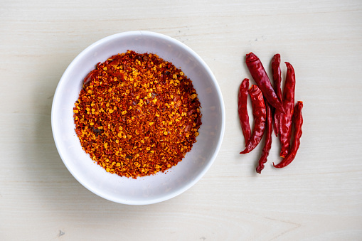 Chilli flakes in a white bowl on a wooden background, with dried red chilies on the side. It is also known as crushed red pepper flakes. Top view.