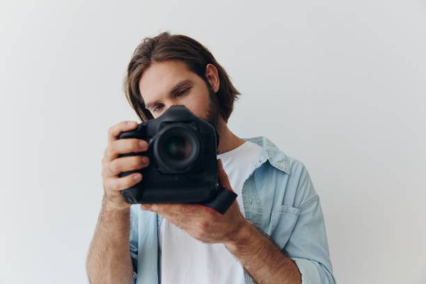 istock Man hipster photographer in a studio against a white background holding a professional camera and setting it up before shooting. Lifestyle work as a freelance photographer 2148785625
