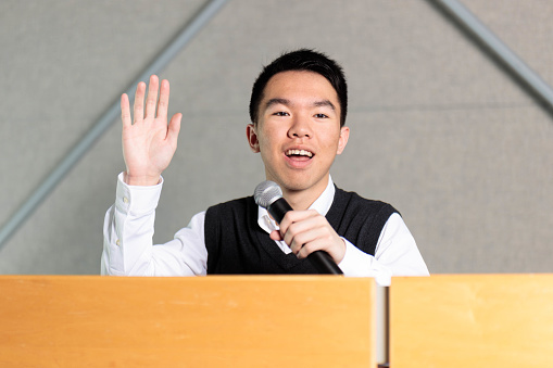 A spokesperson in a dress shirt sits on the stage with a microphone and raises his hand to speak at a formal event
