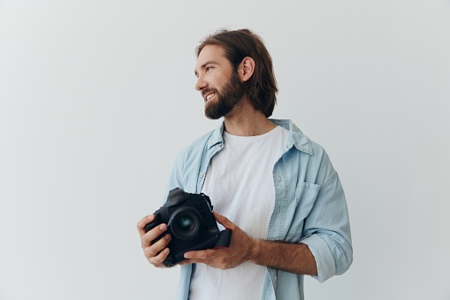 Man hipster photographer in a studio against a white background holding a professional camera and setting it up before shooting. Lifestyle work as a freelance photographer. High quality photo