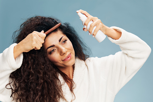 Haircare And Hairstyling. Cheerful Pensive tanned curly Latin lady in bathrobe Spraying On Hair For Repair posing isolated on pastel blue background, using hairbrush. Hair Care Cosmetics Ad Concept