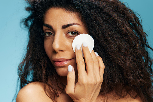 Tanned pretty curly Latin lady holding cotton pad for face, cleansing face with toner after shower over blue studio background. Happy woman enjoying fresh moisturized face skin. Copy space