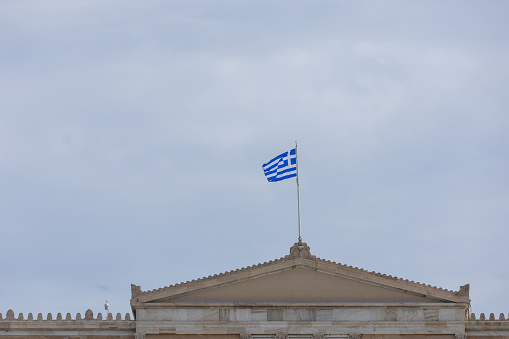 Greek flag on pole over classic building rooftop with blue sky in background