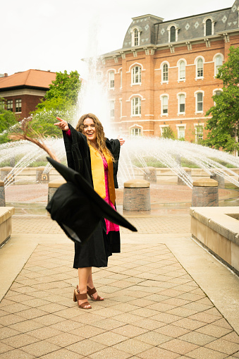 Blond female wearing a mortarboard cap and graduation sash over a pink dress stands at a fountain and throws her mortarboard cap into the air in celebration of graduation day, Indiana, USA